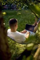 man works on a laptop in a hammock in the garden, next to his pet dog photo