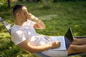 man works on a laptop in a hammock in the garden, next to his pet dog photo