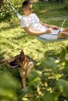 man works on a laptop in a hammock in the garden, next to his pet dog photo
