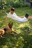 man works on a laptop in a hammock in the garden, next to his pet dog photo