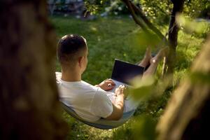 man works on a laptop in a hammock in the garden, next to his pet dog photo