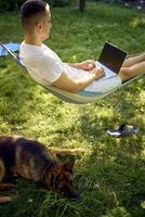 man works on a laptop in a hammock in the garden, next to his pet dog photo