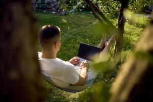 man works on a laptop in a hammock in the garden, next to his pet dog photo
