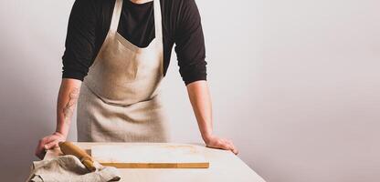 Men cook in a cotton apron holds a wooden rolling pin at the kitchen table with a cutting board. Rustic style. photo