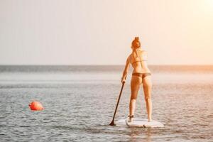 Sea woman sup. Silhouette of happy middle aged tanned woman in rainbow bikini, surfing on SUP board, confident paddling through water surface. Idyllic sunset. Active lifestyle at sea or river. photo