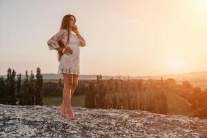 Happy woman in white boho dress on sunset in mountains. Romantic woman with long hair standing with her back on the sunset in nature in summer with open hands. Silhouette. Nature. Sunset. photo