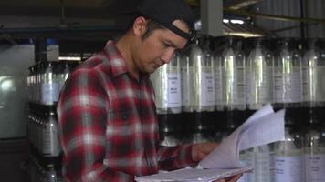 An Asian worker in a red plaid shirt checks beer stocks behind several large beer barrels. video