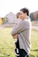 Smiling dad walks with a little girl in his arms through a green meadow. Side view photo