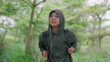 une homme portant des lunettes et une sac à dos portant une vert veste écoute à musique. marcher par une tropical forêt, prendre dans le vues, avoir aventure, et apprécier le tranquillité de la nature video