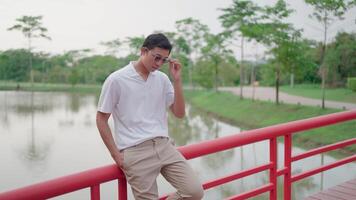 Peace and relaxation amidst nature A man wearing glasses sits on the railing of a red wooden bridge and looks at the nature around him. Take a vacation in a park filled with nature video