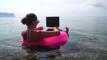 Woman freelancer works on laptop swimming in sea on pink inflatable ring. Happy tourist in sunglasses floating on inflatable donut and working on laptop computer in calm ocean. Remote working anywhere video