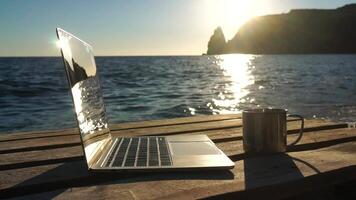 Silhouette of laptop and cup on the beach over sea sunset. Sparkling sea water on background. Freelancer, blogger working on notebook on the beach. video