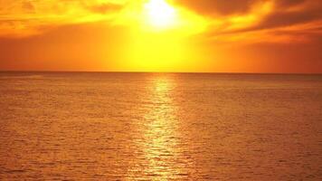 A red burning sunset over sea with rocky volcanic cliff. Abstract nature summer ocean sea background. Small waves on golden warm water surface with bokeh lights from sun. Weather and climate change video