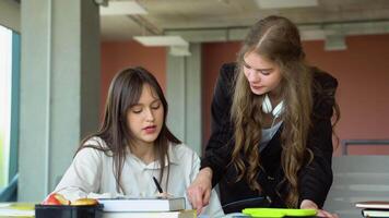 Two female students sitting by desk in lecture hall and discussing passage in book while one of them pointing at page video