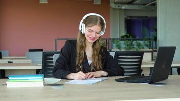 sorridente menina com fone de ouvido estudando on-line, usando computador portátil às universidade, levando notas, cópia de espaço video