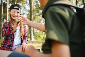 Group of curious happy school kids in casual clothes with backpacks exploring nature and forest together on sunny autumn day, girl holding magnifying glass and looking at fir cone in boy hands. photo