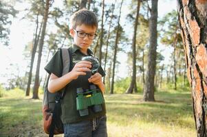 schoolboy is exploring nature with magnifying glass. Little child is looking on leaf of fern with magnifier. Summer vacation for inquisitive kids in forest. Hiking. Boy-scout. photo