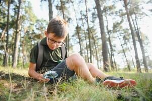 schoolboy is exploring nature with magnifying glass. Summer vacation for inquisitive kids in forest. Hiking. Boy-scout. photo