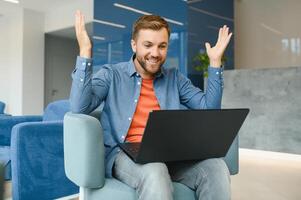 happy smiling remote online working man in casual outfit with laptop in joyful successful winning gesture sitting in an coworking office at a work desk. photo