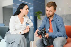 Photographer in office shows photo model in camera. Consultant adjusts activities company as whole. Photo session employees in workplace. Manager well-executed project. Photos best employees company.