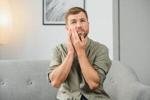 Problems with teeth. Toothache. Upset caucasian guy is sitting on the couch at home, holding his hand to his cheek, having a toothache, needs a dentist consultation, sad facial expression. photo