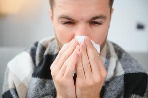 Young man suffering from a common cold and flu or allergy sit at home wrapped in blanket and wipe his nose with tissues while he have a strong headache pain, healthcare concept. photo