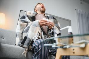 Young man suffering from a common cold and flu or allergy sit at home wrapped in blanket and wipe his nose with tissues while he have a strong headache pain, healthcare concept. photo