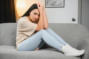Portrait of beautiful young woman with depressed facial expression sitting on grey textile couch. Cyber bullying victim concept. Sad female in her room. Background, copy space photo