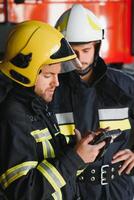 Two professional firefighters with uniforms and protective helmets posing infront of a firetruck. photo
