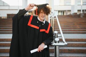 Graduation from university. Young smiling boy university graduate in traditional bonet and mantle standing and holding diploma in hand over university building background photo
