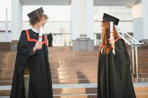 Portrait of happy graduates. Two friends in graduation caps and gowns standing outside university building with other students in background, holding diploma scrolls, and smiling photo