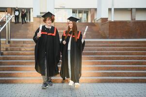 Portrait of happy graduates. Two friends in graduation caps and gowns standing outside university building with other students in background, holding diploma scrolls, and smiling photo