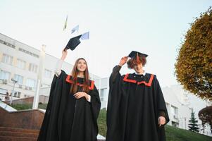 Portrait of happy graduates. Two friends in graduation caps and gowns standing outside university building with other students in background, holding diploma scrolls, and smiling photo