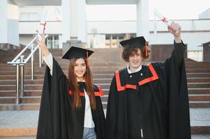 Portrait of happy graduates. Two friends in graduation caps and gowns standing outside university building with other students in background, holding diploma scrolls, and smiling photo