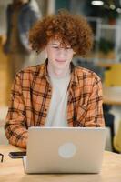 Young happy male freelancer in casual clothes sitting in cafe with laptop and using mobile phone. photo