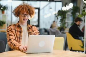 Portrait of Caucasian male freelancer in trendy apparel sitting at cafeteria table and doing remote work for programming design of public website, skilled software developer posing in coworking space. photo