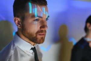 Stock Market Top Trader Looks at Projected Ticker Numbers and Graphs Running, Analysing Data to Make Best Sell. Behind Him Room Full of Screens and Statistics. photo