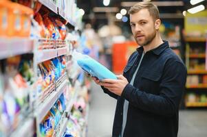 Young man in the supermarket in the household chemicals department. Large selection of products. A brunette in a glasses and a beard in a beige coat. photo