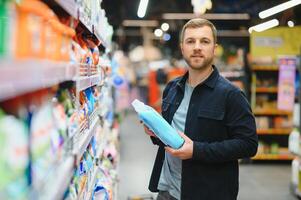 Young man in the supermarket in the household chemicals department. Large selection of products. A brunette in a glasses and a beard in a beige coat. photo
