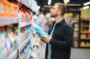 Young man in the supermarket in the household chemicals department. Large selection of products. A brunette in a glasses and a beard in a beige coat. photo