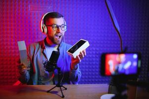 Male technology blogger recording blog or vlog review of new smartphone, showing it on camera at home studio. Blogging, Work from Home concept. Selective focus. Web Banner photo