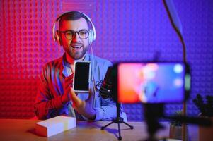 Male technology blogger recording blog or vlog review of new smartphone, showing it on camera at home studio. Blogging, Work from Home concept. Selective focus. Web Banner photo