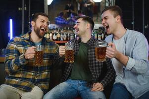 Three young men in casual clothes are smiling and clanging glasses of beer together while sitting at bar counter in pub photo