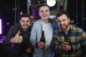 Three young men in casual clothes are smiling, holding bottles of beer while standing near bar counter in pub photo