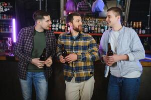 Three young men in casual clothes are smiling, holding bottles of beer while standing near bar counter in pub photo