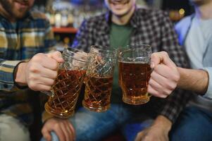 Three young men in casual clothes are smiling and clanging glasses of beer together while sitting at bar counter in pub photo