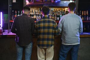 Three young men in casual clothes are smiling, holding bottles of beer while standing near bar counter in pub photo