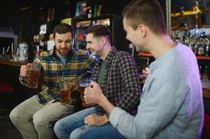 Three young men in casual clothes are smiling, holding bottles of beer while standing near bar counter in pub photo