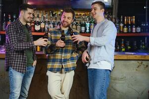 Three young men in casual clothes are smiling, holding bottles of beer while standing near bar counter in pub photo