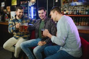 Three young men in casual clothes are smiling, holding bottles of beer while standing near bar counter in pub photo